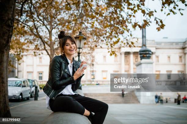秋のシーズン、ロンドンのトラファルガー広場でスマート フォンを使用して幸せな女 - レザージャケット ストックフォトと画像