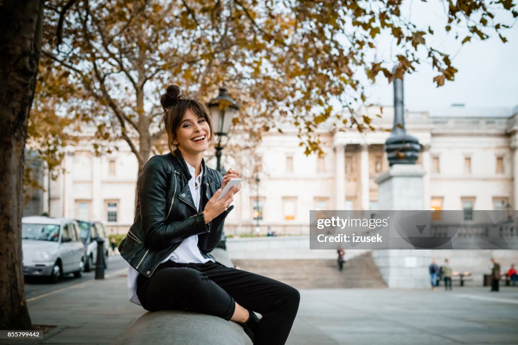 Happy woman using smart phone at Trafalgar Square in London, autumn season