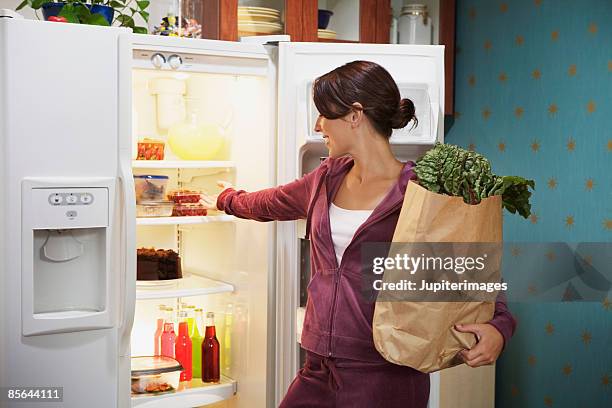 woman looking in refrigerator and holding grocery bag - sportswear shopping stock-fotos und bilder