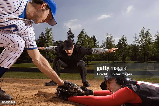 baseman trying to strike out baseball player while umpire signals him safe - baseball umpire fotografías e imágenes de stock