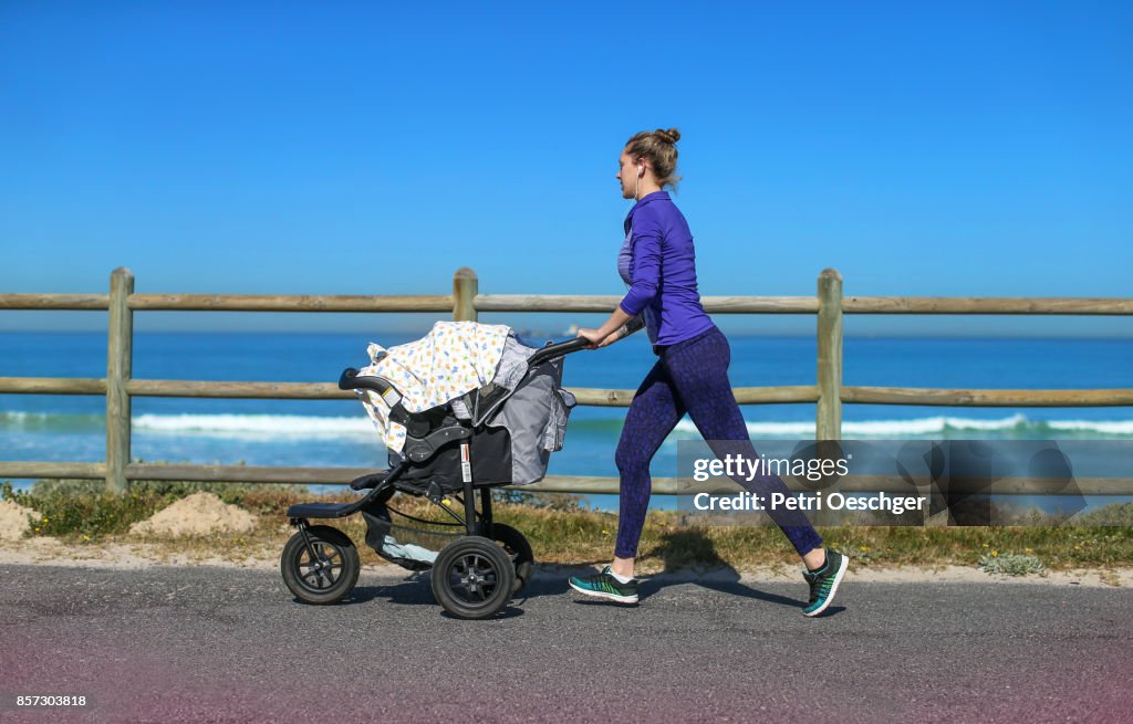 A Young mother jogging on the beachfront pushing a stroller.