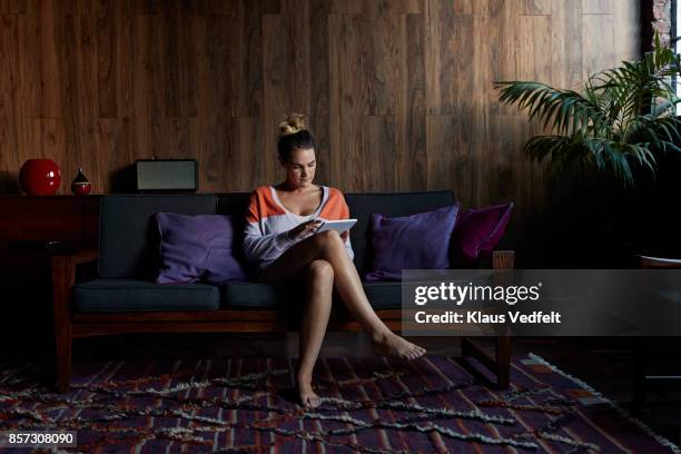 young woman sitting in couch and using digital tablet - de pernas cruzadas imagens e fotografias de stock