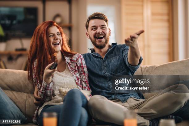 young couple laughing while watching tv at home. - young couple watching tv stock pictures, royalty-free photos & images