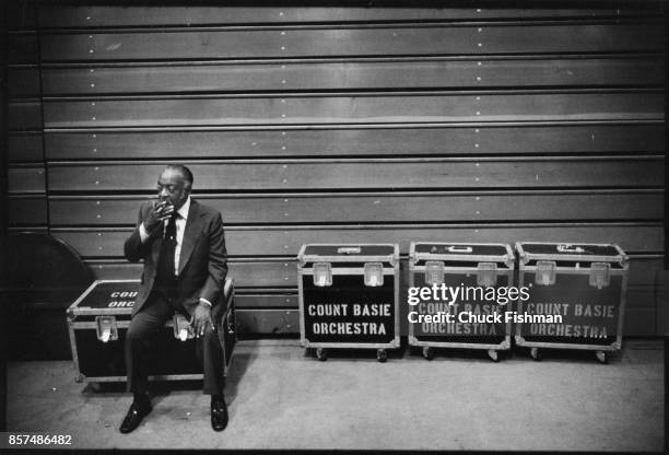 American Jazz and Swing musician Count Basie smokes a cigar as he sits on a trunk backstage in a college gymnasium before a performace, August 1978.