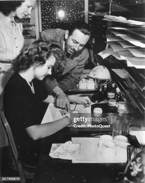 American movie producer, artist, and animator Walt Disney leans over painter Edith Moore as she works on an animation cel , Burbank, California, 1943.