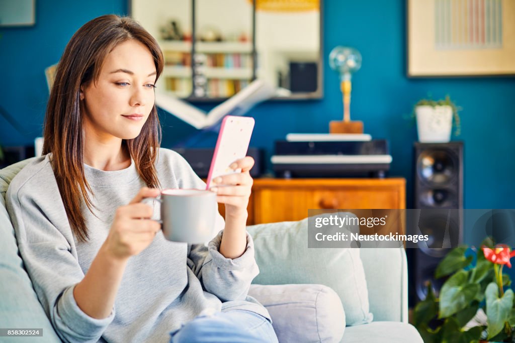 Beautiful woman using smart phone and having coffee on sofa