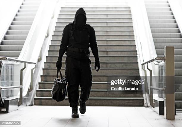 hooded lone wolf man wearing black carrying bag in urban underground public transport setting - terrorismo imagens e fotografias de stock