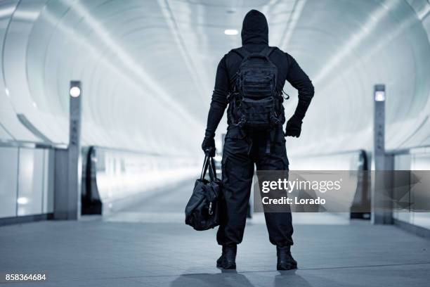 hooded lone wolf man wearing black carrying bag in urban underground public transport setting - terrorism stock pictures, royalty-free photos & images