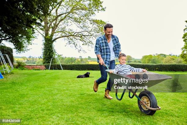 man giving son ride in wheelbarrow - family garden play area stock pictures, royalty-free photos & images