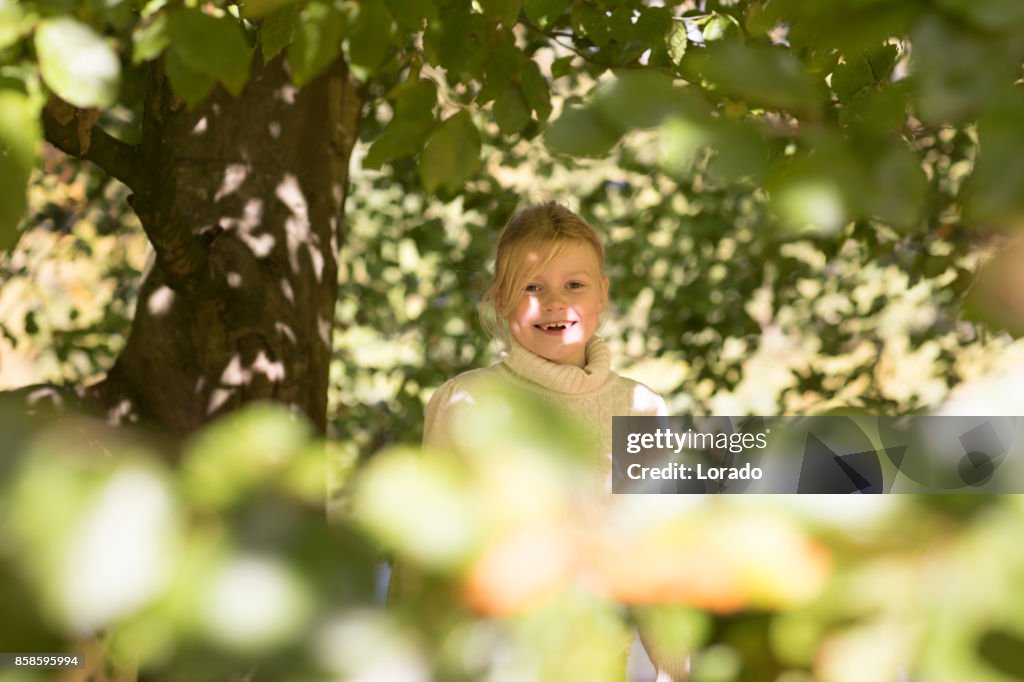 Beautiful young blond girl playing in trees in Autumnal Park