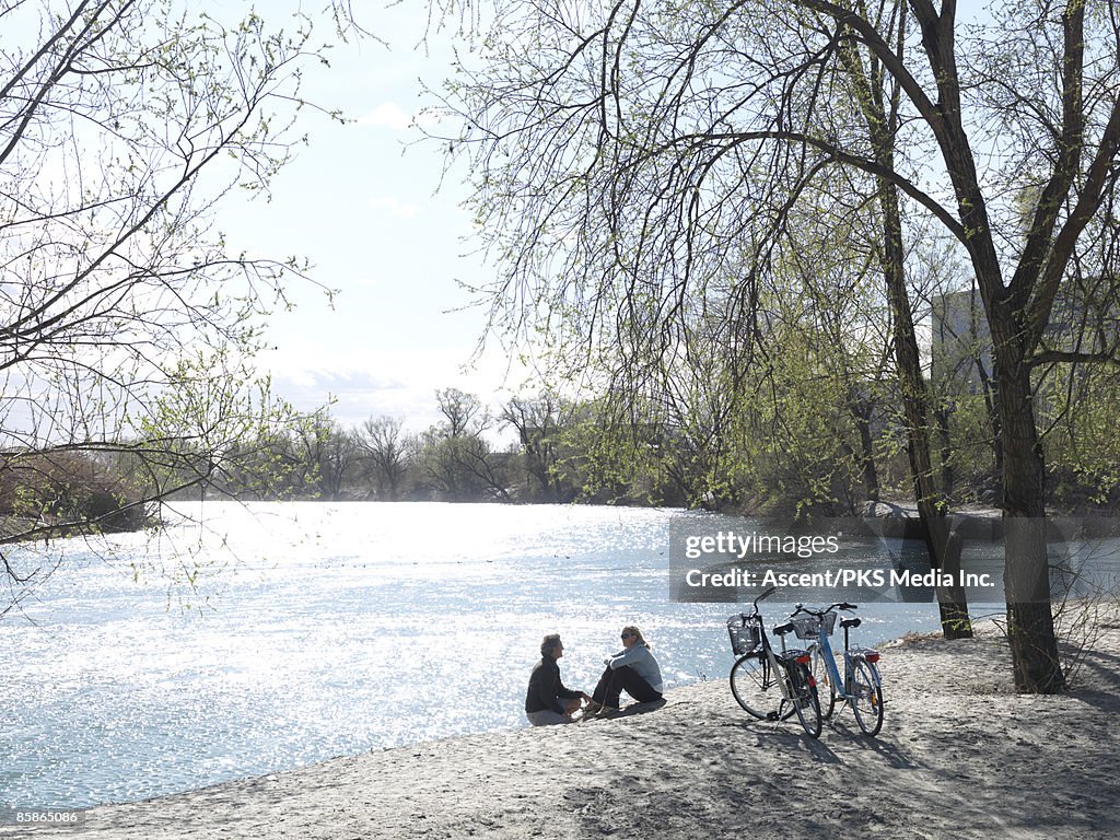 Couple with bikes sit at edge of river, talking