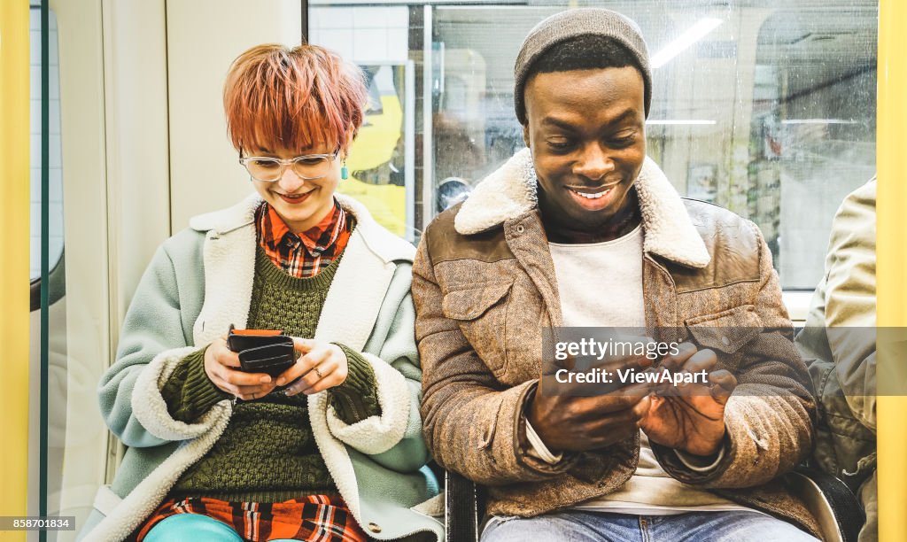 Multiracial hipster friends couple having fun with smartphone in subway train - Urban relationship concept with young people watching mobile phone in city underground area - Bright desaturated filter