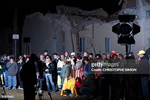 Quake survivors and volunteers watch a news programme on a television set placed on the ground, as a journalist waits for a live broadcast at the...