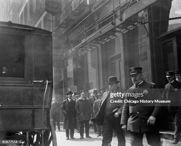 Reputed gangster James Franche and a policeman walking across a sidewalk away from a building with other policemen standing in the entrance, toward...