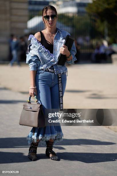 Guest wears sunglasses, a blue ruffle denim jacket, blue ruffle pants jeans, a Hermes bag, a "Girl Gang" belt outside Nina Ricci, during Paris...