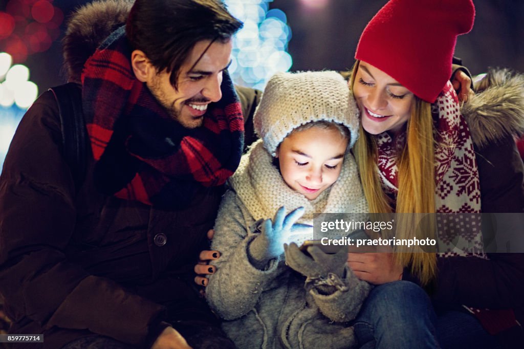 Joven, pareja feliz es abrazando a su hija hasta que ella está enviando mensajes de texto en Navidad