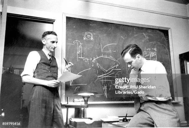 Candid of two men in a Johns Hopkins University classroom in front of a blackboard covered in mathematical equations, including James Herbert Henry,...