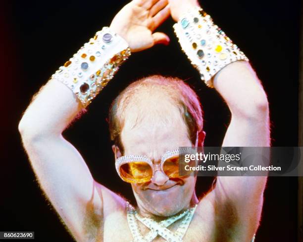 Close-up of English musician Elton John, shirtless but for several sequinned straps and wristbands, as he performs, arms above his head, 1970s.