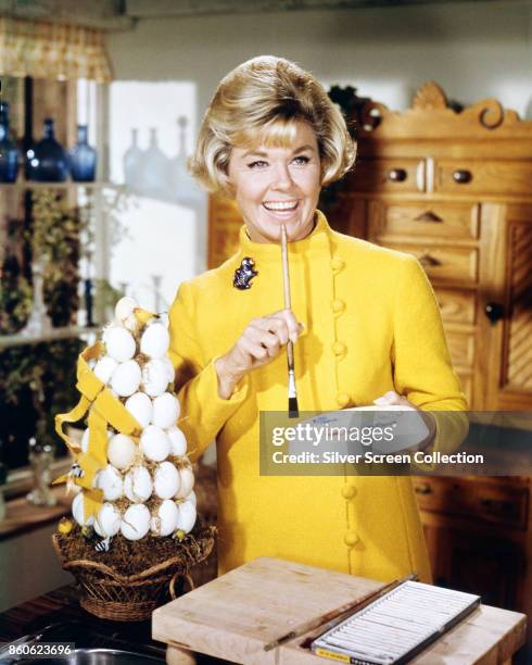 Portrait of American actress Doris Day, dressed in a high-neck, yellow jacket with a brooch on her right shoulder, as she prepares to paint a basket...