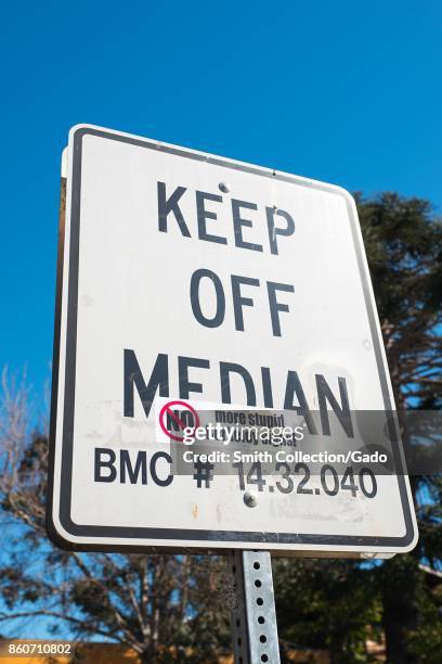 Road sign reading "Keep Off Median", on which someone has placed a sarcastic decal reading "No More Stupid Berkeley Signs", on a road median which is...