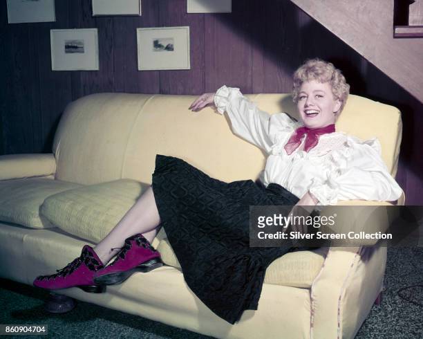 Portrait of American actress Shelley Winters as she laughs, seated on a sofa, 1950s or 1960s. She wears a red scarf, a white blouse with lace and...