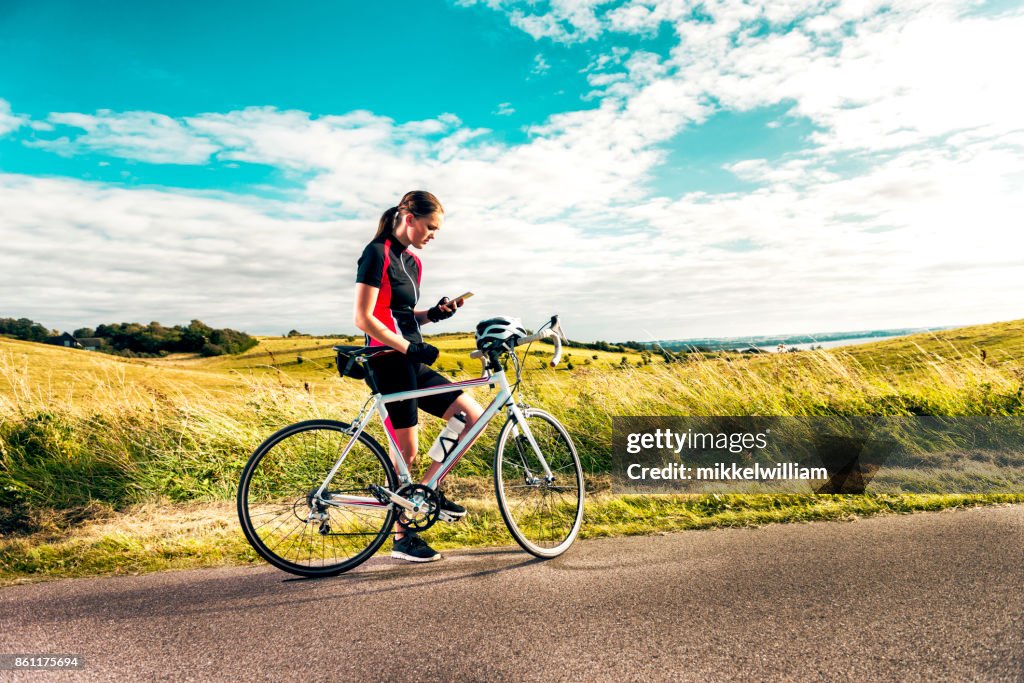 Sporty woman on racing bicycle uses mobile phone while exercising on country road