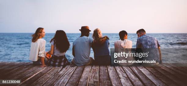 young multi-ethnic couples and friends sitting on wooden jetty together - beach pier stock pictures, royalty-free photos & images