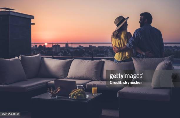 vista traseira do casal feliz se comunicam em uma varanda de cobertura ao nascer do sol. - apartamento de cobertura - fotografias e filmes do acervo