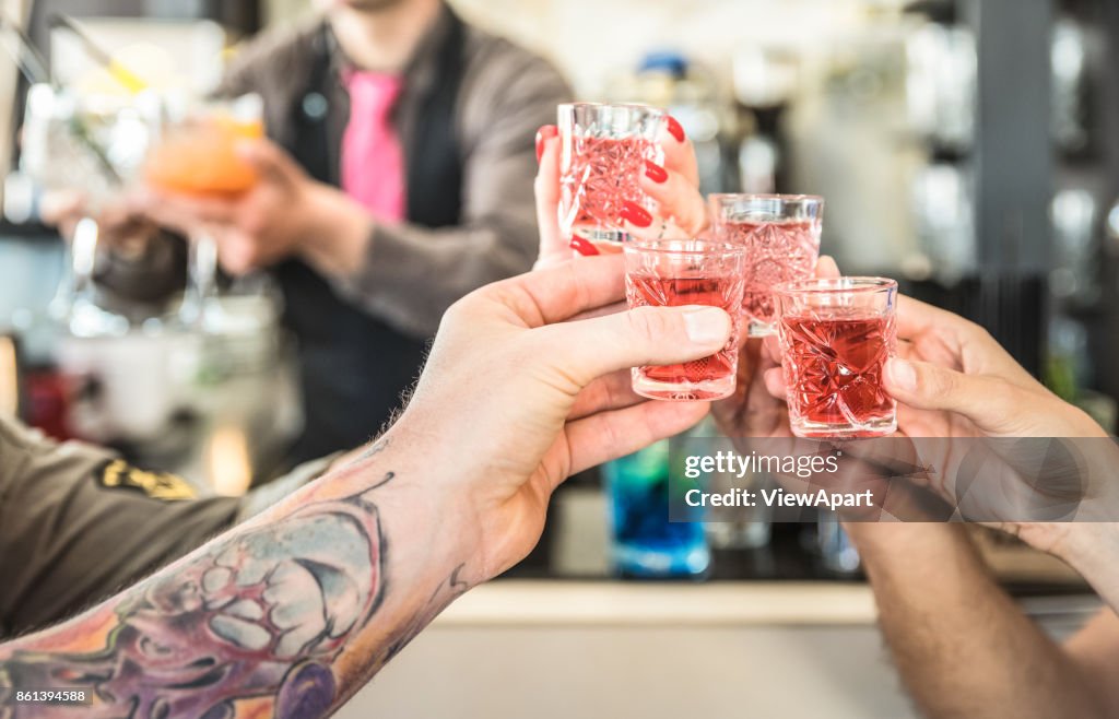 Group of drunk friends toasting cocktails at bar restautant - Food and beverage concept on nightlife moments - Defocused bartender serving drinks on background - Focus on hands cheering red shot glass