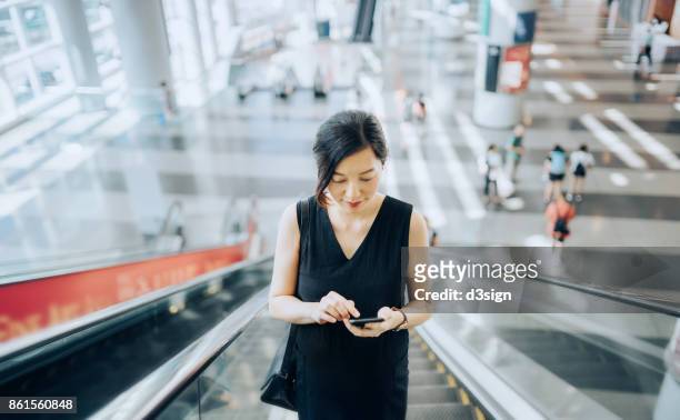 young businesswoman reading emails on smartphone while riding on escalator - hauptverkehrszeit stock-fotos und bilder