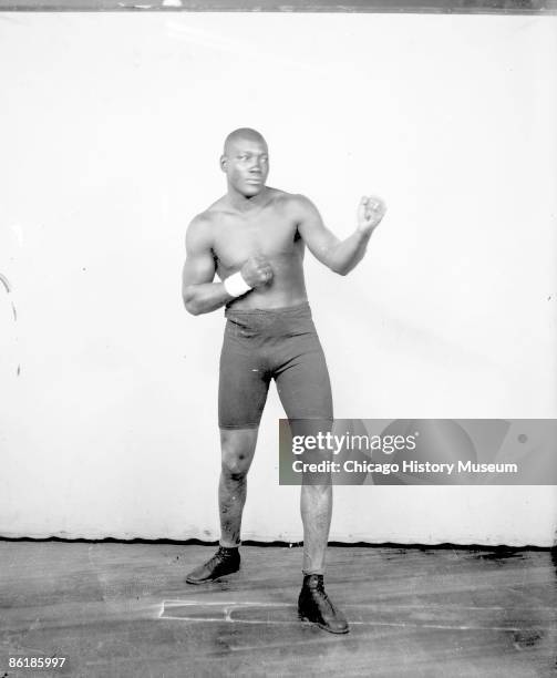 Portrait of Jack Johnson, African American heavyweight champion boxer, standing in an ordinary boxing stance in front of a light colored background...