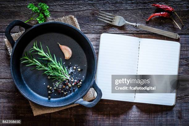 cookbook and cast iron pan with some herbs on wooden table - cookbook stock pictures, royalty-free photos & images