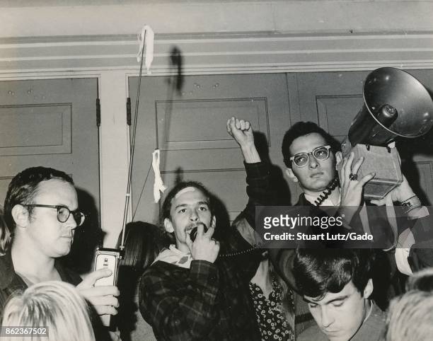 Student wearing hippie attire holding his fist into the air and speaking into the microphone of a bullhorn loudspeaker during an anti Vietnam War...