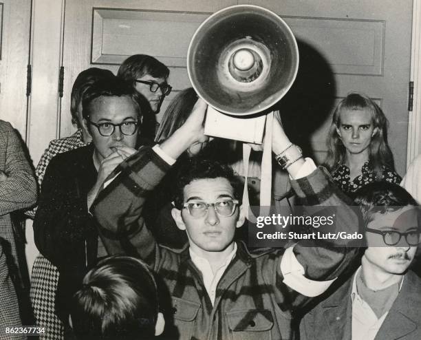 Student wearing hippie attire holds a bullhorn loudspeaker above his head while another student speaks into the microphone during an anti Vietnam War...