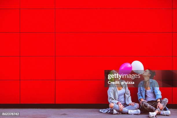 due giovani amiche che si divertono, facendo esplodere palloncini davanti al muro rosso - quirky kissing foto e immagini stock