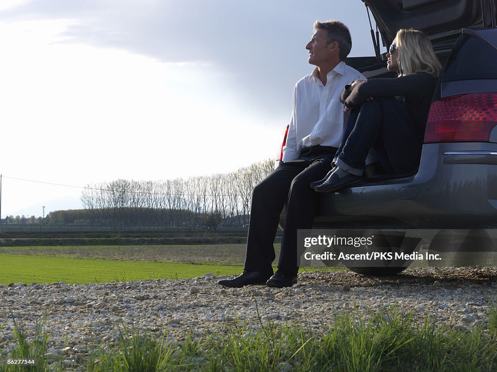 Couple sit on car tailgate,gaze across green field