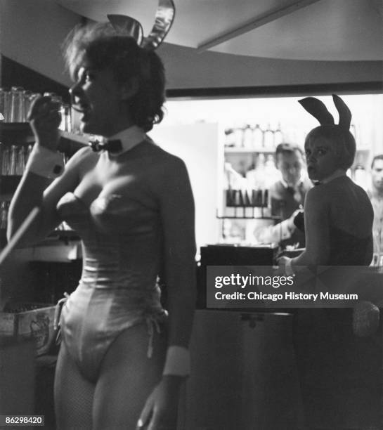 Two Playboy bunnies at work at the Playboy Club, one is seen having a snack, while the other waits for an order at the bar, Chicago, ca.1960s. Two...