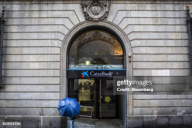 Pedestrian carries a blue umbrella past the entrance to a Banco de Sabadell SA bank branch on Sant Jaume square in Barcelona, Spain, on Thursday,...