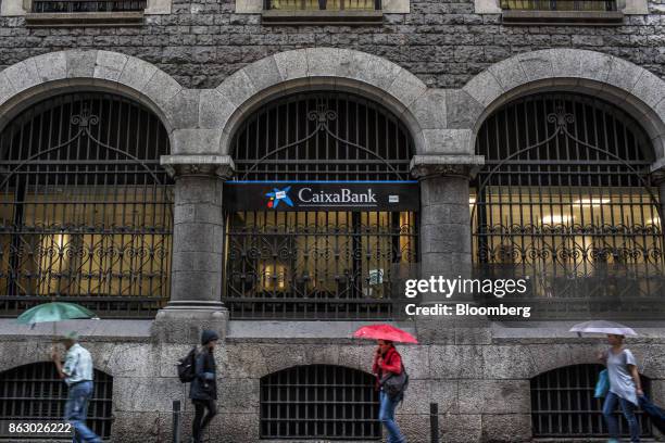 Pedestrians walk beneath umbrellas past a CaixaBank SA bank branch on Via Laietana in Barcelona, Spain, on Thursday, Oct. 19, 2017. Spains...