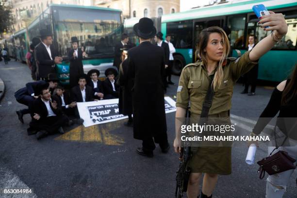 Female Israeli soldier uses her cell phone to take a selfie photograph with ultra-Orthodox Jewish demonstrators in the background who are impeding...