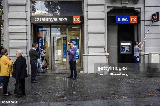 Customers wait to withdraw cash from a Catalunya Bank SA bank branch as a worker cleans a sign above a Banco Bilbao Vizcaya Argentaria SA automated...