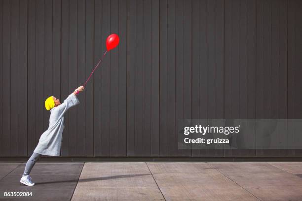 joven bailando y sosteniendo globo rojo contra la pared gris - excéntrico fotografías e imágenes de stock