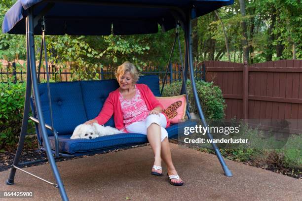 smiling blond caucasian woman in her 50's sits next to her little white ocherese dog on a blue porch swing in the backyard on a summer day, indiana, usa - swing chair stock pictures, royalty-free photos & images