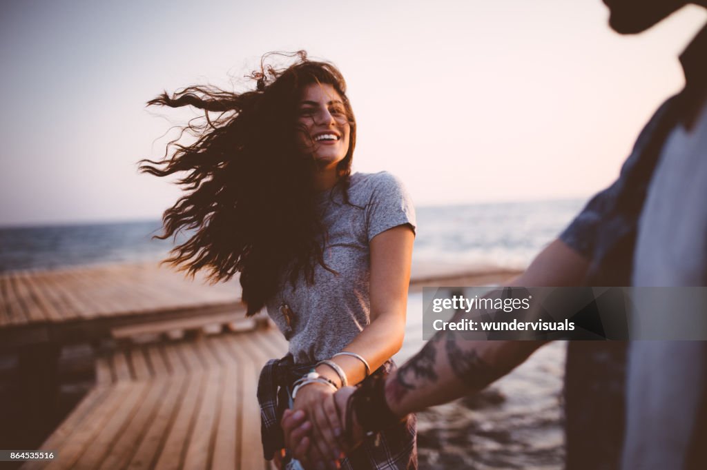 Young playful hipster couple holding hands on jetty at sunset