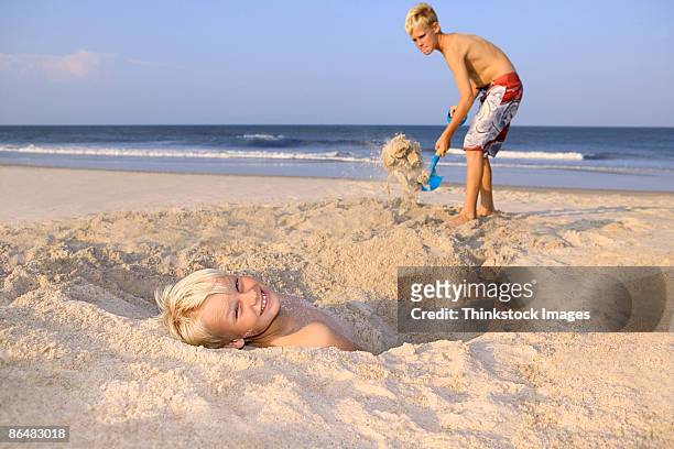 boy burying other boy in sand - digging beach photos et images de collection