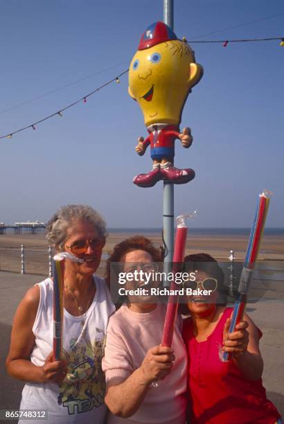 Three laughing ladies hold up their sticks of rock beneath a seaside character on the seafront at Blackpool, on 18th July 1993, Blackpool,...