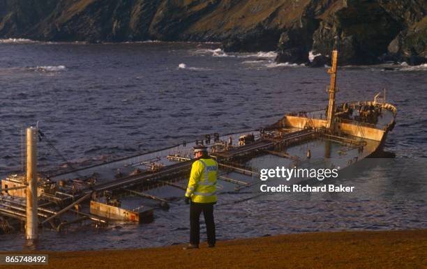 Police officer looks over stormy waves wich crash over the super-structure and funnel of the Liberian-registered MV Braer oil tanker, spilling 84,700...