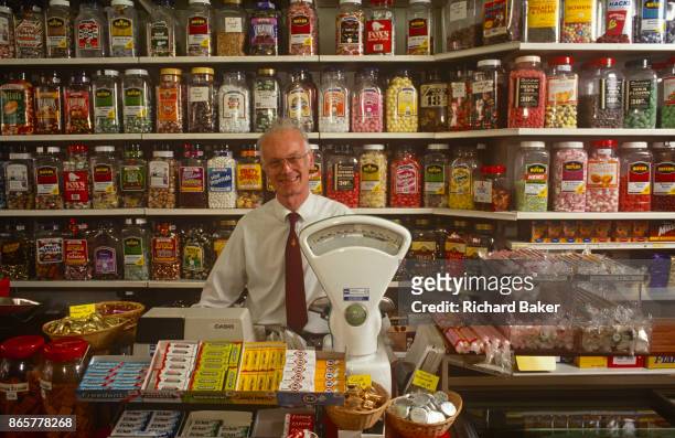 Portrait of a smiling 1990s shop keeper in his sweet store surrounded by jars of confectionery and his traditional pre-digital weighing scales, on...