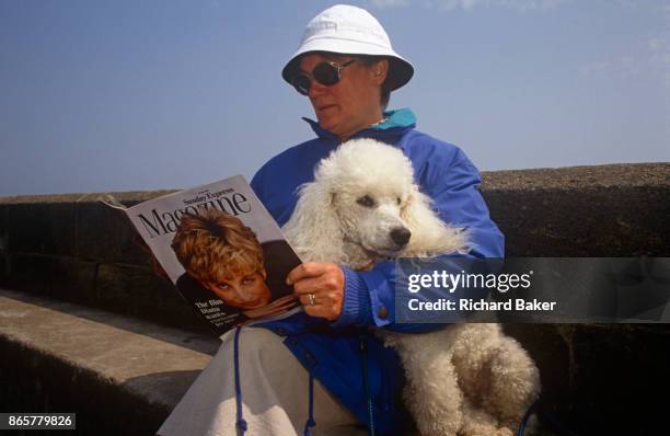 1990s lady sits with her pet poodle on a sea wall reading the Sunday Express magazine that features Princess of Wales on the cover, on 19th July...