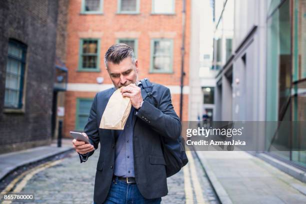 businessman eating a sandwich and using smart phone - eating on the move stock pictures, royalty-free photos & images